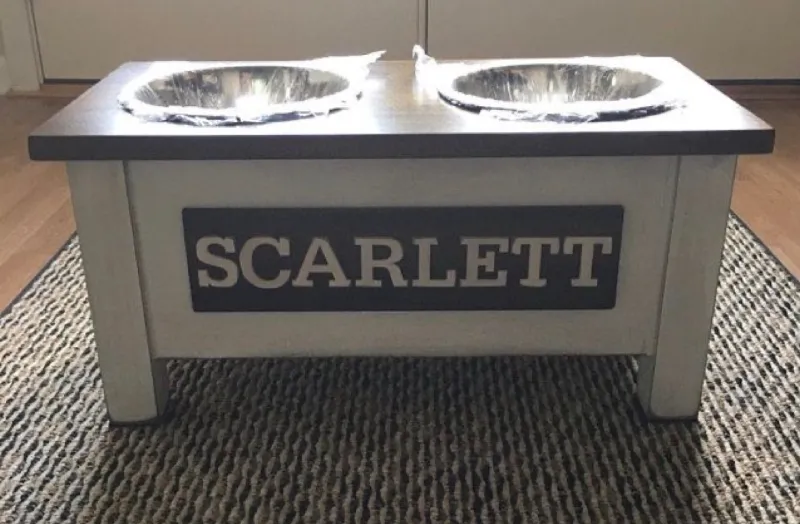 White dog bowl stand with a dark top and a dark nameplate reading Scarlett in white letters, sitting on a woven rug
